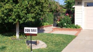 A for-sale sign in front of a home shaded by a mature tree